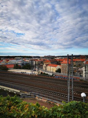 Parçalı bulutlu bir gökyüzü altında renkli binalar ve yeşilliklerle dolu kentsel bir arazide bir demiryolu hattı uzanır. Sahne, şehir hayatının özünü yakalar..