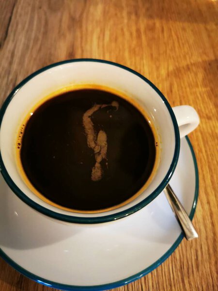 A white cup filled with dark coffee is placed on a rustic wooden table. A metallic spoon is adjacent, enhancing the cozy atmosphere of the setting.