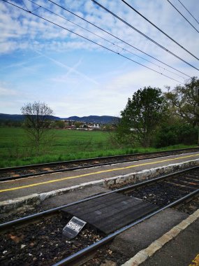 Serin bir tren istasyonu yemyeşil tarlaların ve yumuşak uzak tepelerin olduğu bir zemin üzerine kurulur. Parçalı bulutlu bir gökyüzü bahar boyunca huzurlu bir atmosfer yaratır..