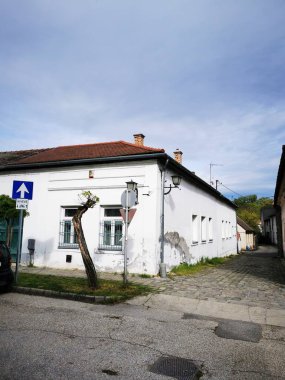 Dar bir caddenin kesiştiği yerde, bulutlu bir gökyüzünün altında, kırsal mimariyle çevrili, barışçıl bir atmosferi yansıtan küçük beyaz bir bina duruyor..