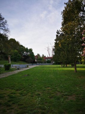 Huzurlu bir park, ağaçlarla çevrili geniş yeşil bir çimenliğe sahiptir. Açık bir yolu vardır. Yumuşak bir öğleden sonra gökyüzünün altında sakin yürüyüşler düzenler..