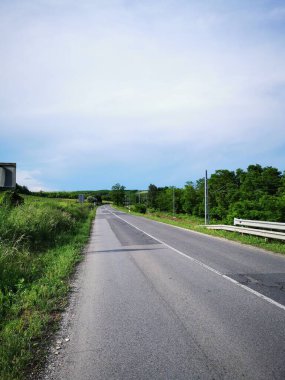 Yol ileride, berrak bir gökyüzünün altında yemyeşil bir kanatla çevrili, gündüz yolculuğu sırasında kırsal yaşamın sükunetini gösteriyor..