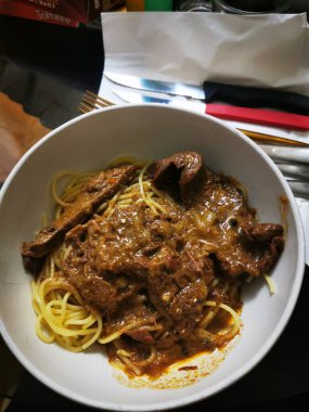 Cömert bir spagetti servisi leziz bir sosta yumuşak et parçalarıyla kaplıdır. Yemek masasına konur ve yanında da basit aletler bulunur..
