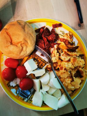 Sağlıklı bir kahvaltı, çırpılmış yumurta, olgun domates, peynir parçaları ve renkli bir tabağa dizilmiş yumuşak bir çöreğe sahiptir. Güne sıcak bir başlangıç için davet eder..