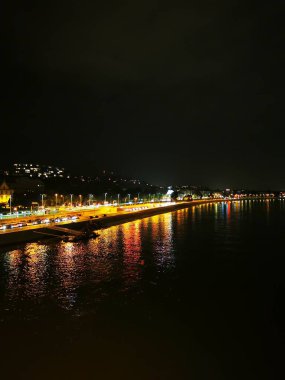 Sakin nehir, taşıtlar hareketli sahil boyunca ilerlerken renkli şehir ışıklarını yansıtır. Sakin atmosfer, deniz kenarındaki kentsel gece hayatını gözler önüne seriyor..