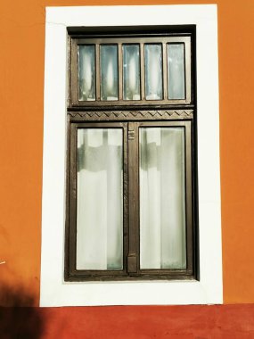 Zarif ahşap pencere, parlak turuncu bir duvara dayalı birden fazla pencere ve dekoratif bir kapak içeriyor, büyüleyici ve davetkar bir atmosfer yaratıyor..