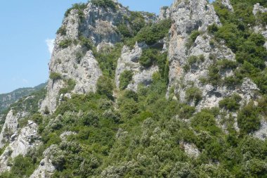 Yükselen uçurumlar parlak yeşilliklerle süslenir, parlak mavi gökyüzüne karşı yükselir, parlak güneş ışığı altındaki uzak dağlık bölgenin güzelliğini gözler önüne serer..