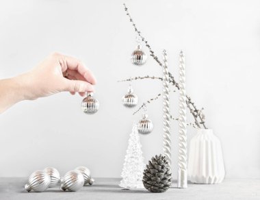 Gri arka planda minimalist noel seti. Gümüş mumlar, noel baloları ve çam kozalakları. Kadın eli süs için topu tutuyor. Kış tatili için İskandinav hipster ev dekorasyonu.