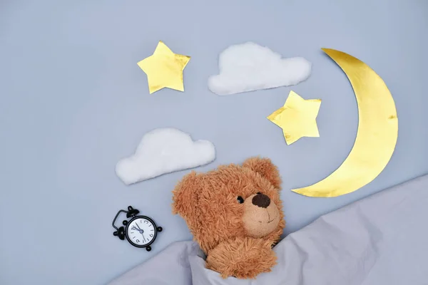 teddy bear sleeping under blanket on grey background. moon, stars and alarm clock. kids healthy sleep concept. cyrcadian rythms and good night time for children