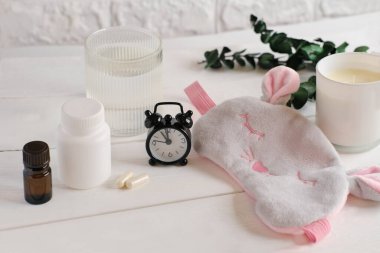 Komodinin üzerinde melatonin kapsülleri ve lavanta yağı ve uyku maskesi. Sağlıklı bir uyku için çalar saat ve kokulu mum. İyi uyku takviyesi. Gece olmadan rahatla..