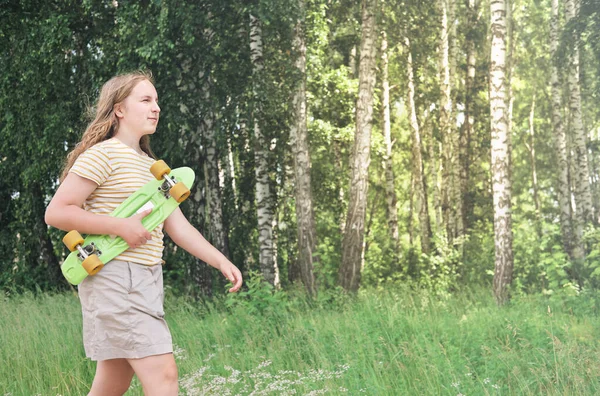 Teenager girl holding a skateboard and going for a skating in a park. blond curly teen girl with long hair enjoiying walking alone — Stock Photo, Image