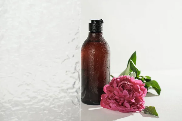 Гель для душа в янтарной бутылке с капельками воды. Стеклянная дверь ванной комнаты и розовый пионский цветок рядом с косметическим небрендированным продуктом в коричневой бутылке. — стоковое фото