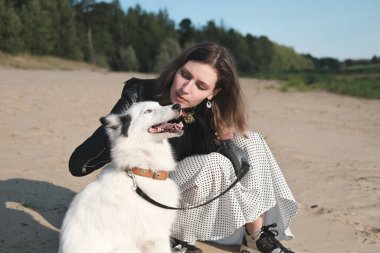 Mutlu laika köpeği gülümsüyor ve sahibinin yanında oturuyor. Genç ve güzel bir kadın kumlu bir sahilde köpeğini okşuyor. Birliktelik ve sevgi dolu evcil hayvanlar konsepti.
