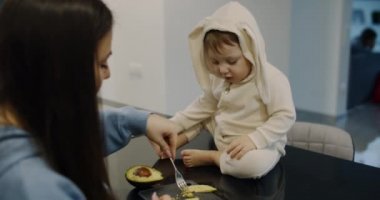 A happy little child, fed with mother's avocado on the kitchen table. Slow.