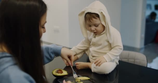 A happy little child, fed with mother's avocado on the kitchen table. Slow. — Vídeo de Stock