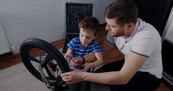 Молодой отец помогает сыну починить велосипед инструментами в доме. — стоковое видео