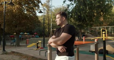 A thoughtful man stands quietly in a park, reflecting deeply among vibrant fall foliage and playground equipment