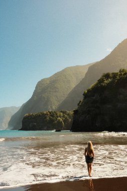 Gözlerden uzak kıyı yürüyüşü, okyanus manzaralı sahil yürüyüşü, güneşli gökyüzünün altında sakin kıyı şeridi boyunca gezinen yalnız bir kadın.