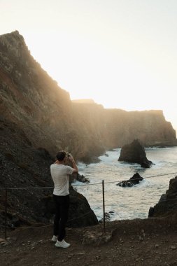 Denizci gün batımını yakalar, peyzaj sanatçısı vahşi dalgaları ve sakin günbatımı renklerini vurgulayan kıyı manzarasını çeker.