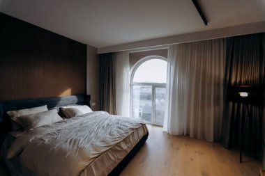 Karanlık çöktüğünde karamsar yatak odası, çevre aydınlatması ve kemerli pencere silüeti. Derin gölgeler, sıcak lamba, katmanlı tekstil, samimi.