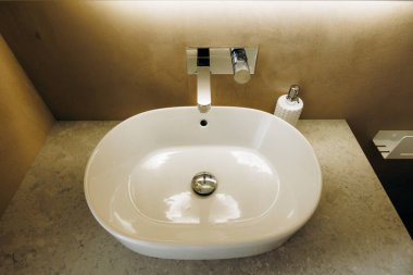 Minimalist beyaz oval lavabo taş tezgahın üzerinde krom musluk, porselen leğen, yakınlarda sabun makinesi, sıcak ortam aydınlatması,