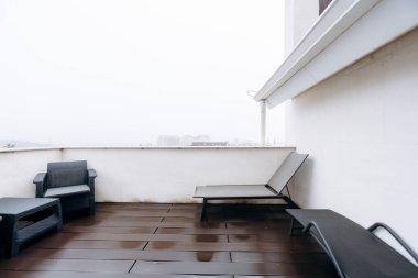 Boş, ıslak teras güvertesi, beyaz duvara karşı modern koltuklar, son yağmurdan parlayan koyu ahşap kalaslar, alçak sis.