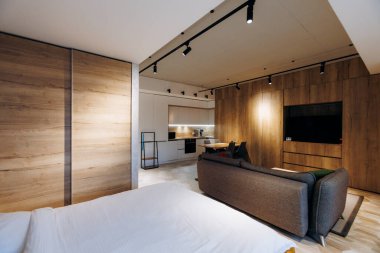 Çağdaş stüdyo dairesi ahşap aksanlı ve açık planlı yatak, kanepe, mutfak, duvara monte edilmiş televizyon.,