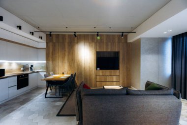 Kanepesi, ahşap aksanlı duvarı, entegre mutfağı, sıcak ortam aydınlatması ve günlük yemek masası olan açık planlı oturma odası.