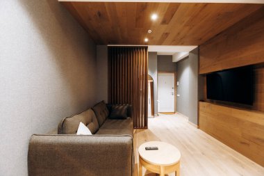 Ahşap tavanlı ve panelli rahat salon koridoru, kompakt kanepe ve tabure, duvara monte edilmiş televizyon, ince sıralı aydınlatma,