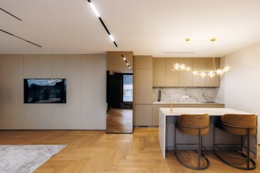 Mutfak adası ve duvara monte edilmiş televizyonu, uyumlu nötr paleti, parke döşemesi ve katmanlı katmanı olan açık planlı yaşam alanı.