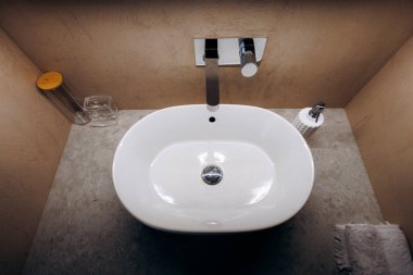 Taş tezgahta oval beyaz lavabo, modern krom musluk, sıcak bej alçı duvarlar, temiz cam bardak ve sabun tabağı, ince ince