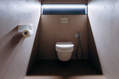 LED rafı olan minimum tuvalet oyuğu. Temiz porselen kase yatay aksan ışığının altında, bidet hortumu görünür, kompakt