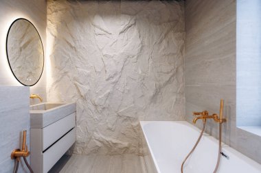 Kalıplı taş duvara karşı serbest küvet sıcak pirinçten bir musluk. Aynadan aydınlatılmış doğal aydınlık ahşap kaplıca.