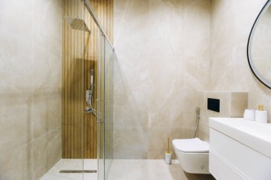 Cam duş ve bej mermer duvarlar dingin spa havası yaratıyor, duvara monte edilmiş tuvalet, yağmur ve el yapımı duş eşyalar, dikey