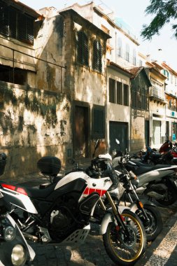 Eski model motorlar güneş ışığı altında bekliyor, eski yıpranmış binanın yanına park edilmiş çeşitli klasik motosikletler koleksiyonu.