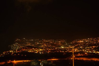 Yoğun tepe yamacı, şehir ışıklarıyla parıldıyor. Geniş gece manzarası, şehir enerjisini ve ışıltısını taşıyor.