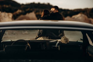 Çiftler, açık havada mahrem bir anı paylaşır, günbatımı parıltısı, klasik araba çatı sahnesini kucaklayan çifti aydınlatır.