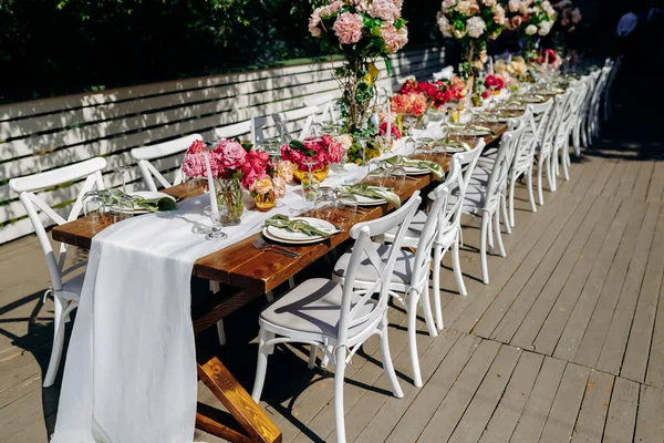 Açık hava kutlaması, güneşli açık hava ziyafet masası yemyeşil çiçekler ve rafine yemek aksesuarlarıyla süslenmiş.