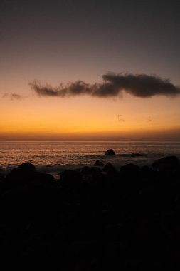Canlı günbatımı sahnesi, kızgın gökyüzünün altında siluetli kayalar, gölgeli taşlar ve parlayan ufuklarla yoğun günbatımı.