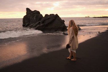 Akşam kıyılarında gezinen kadın, sandaletli ve süveterli kadın, ıslak kumlarda düşünceli yalnız gezintiye çıkar.