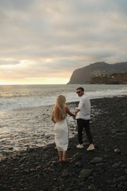 Çift, altın saat boyunca kayalık sahilde ışık anı paylaşır, güler yüzlü etkileşimler, günlük kıyafetler ve sıcak sahil ışıltısı