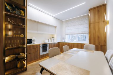 Beyaz masası ve meşe kabini olan parlak yemek alanı, LED striptiz ışıkları, raflarda sergilenen cam döşeme, açık tasarım