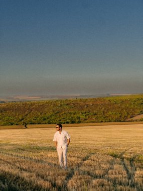 Gün batımında buğday tarlasında yürüyen adam, günlük beyaz kıyafet, uzak yuvarlanan tepeler, sıcak alçak ışık uzun gölgeler, eller