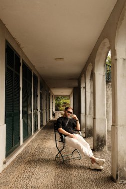 Kemerli koridorda oturan adam, rahat bir poz, sıcak ışık altında, klasik panjurlar, yürüyüş yolu, desenli fayanslı döşeme, gündelik çarşaf