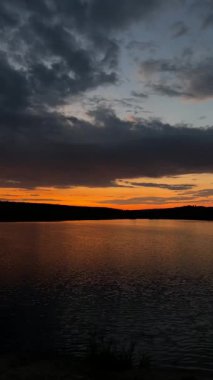 Durgun göl üzerinde alacakaranlık ufuk, ince turuncu şeritli derin deniz bulutları, cam gibi su yansıtan gökyüzü, uzak ağaç çizgisi silueti.