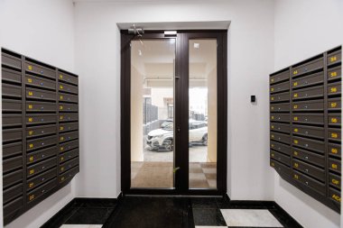 Posta kutuları, camdan kapı girişleri görünür arabaların olduğu otopark manzarası, çift kapılı şeffaflık, güvenlik tuş takımı.