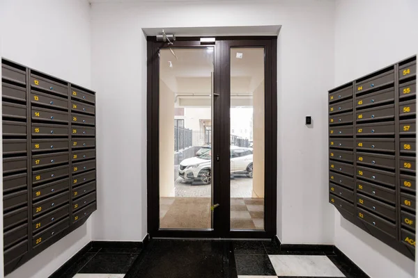 Posta kutuları, camdan kapı girişleri görünür arabaların olduğu otopark manzarası, çift kapılı şeffaflık, güvenlik tuş takımı.