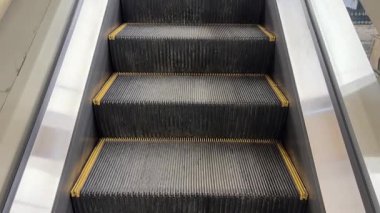 Kapalı yürüyen merdiven, metal hareketler, desenli lastik izleri ve sarı emniyet kenarları olan boş alışveriş merkezi geçiş sahnesi, cam balüstrad.