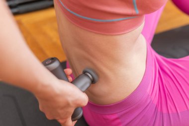 Genç bir kadın vücudun ve kasların ağrısını dindirmek ve rahatlamak için perküsyon masaj tabancası kullanıyor. 