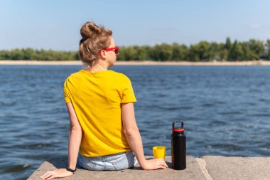 Genç bir kadın rıhtımda oturmuş, yazın güneşli bir günde elinde büyük bir termos ve bardakla mavi denizin manzarasını seyrediyor.